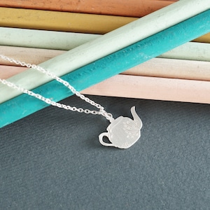 Puede incluir: Un collar de plata con un colgante de tetera. La tetera está decorada con un diseño floral.