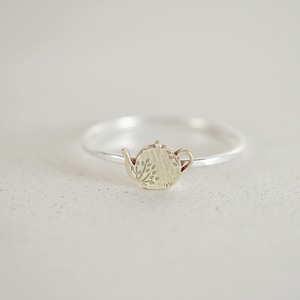 Könnte beinhalten: Ein silberner Ring mit einem kleinen, goldenen Teekannen-Anhänger. Die Teekanne hat ein detailliertes Design mit Blättern und eine strukturierte Oberfläche.