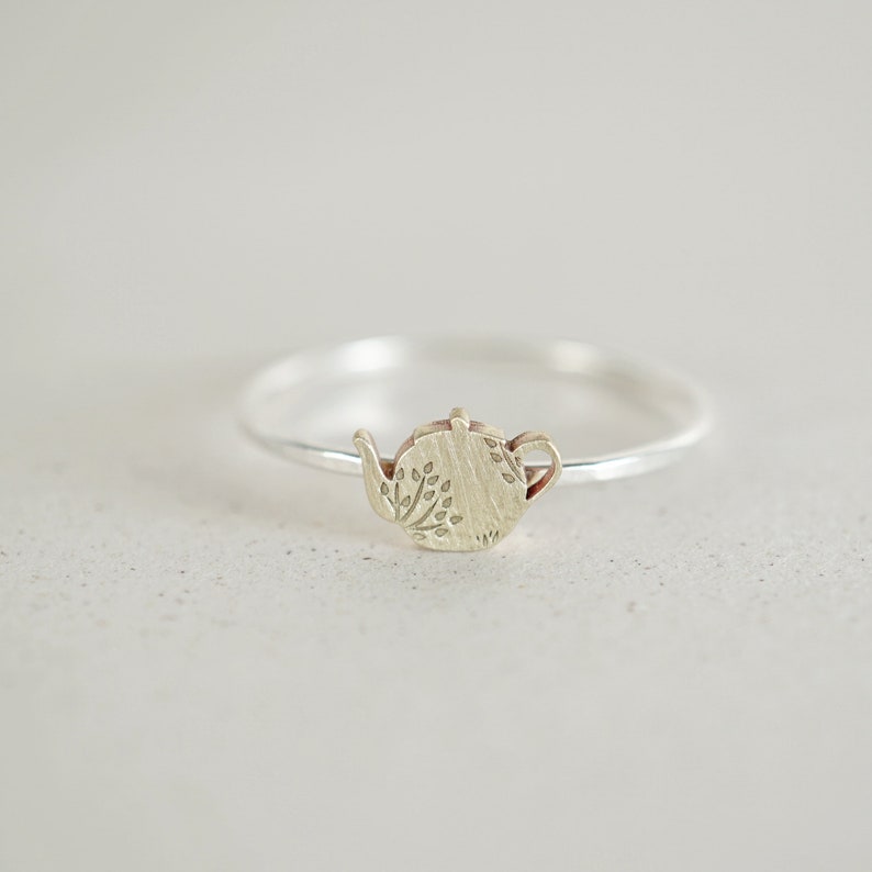 K&ouml;nnte beinhalten: Ein silberner Ring mit einem kleinen, goldenen Teekannen-Anh&auml;nger. Die Teekanne hat ein detailliertes Design mit Bl&auml;ttern und eine strukturierte Oberfl&auml;che.
