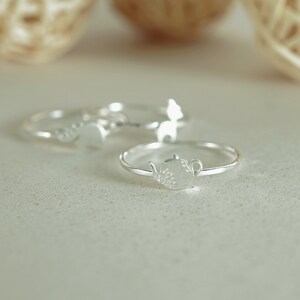 K&ouml;nnte beinhalten: Drei silberne Ringe mit unterschiedlichen Designs. Der Ring im Vordergrund zeigt eine Teekanne mit einem floralen Design.