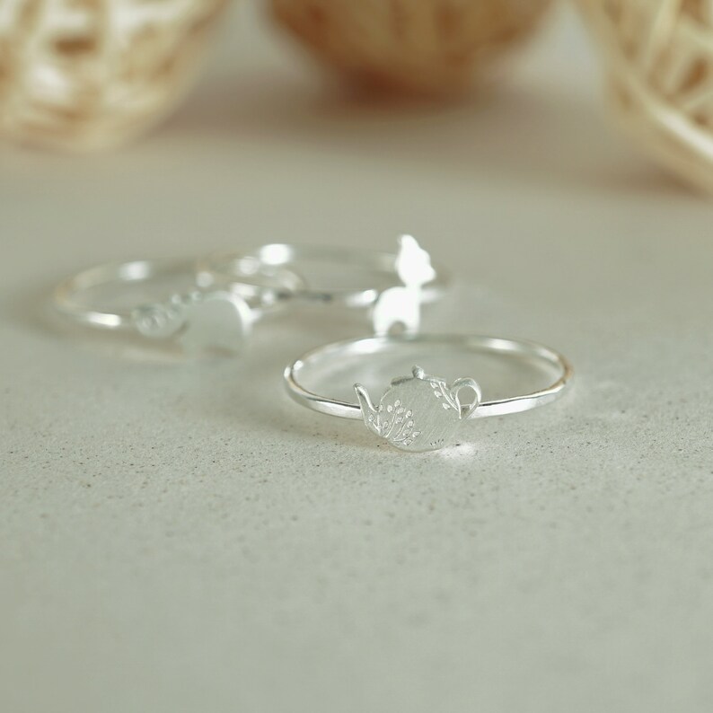K&ouml;nnte beinhalten: Drei silberne Ringe mit unterschiedlichen Designs. Der Ring im Vordergrund zeigt eine Teekanne mit einem floralen Design.