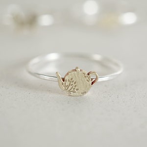 K&ouml;nnte beinhalten: Ein silberner Ring mit einem Teekannen-Anh&auml;nger. Die Teekanne ist mit einem floralen Muster verziert.