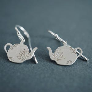 Op de afbeelding: Een paar zilveren oorhangers met een theepotontwerp met een bloemenpatroon. De oorhangers zijn gemaakt van sterling zilver en hebben een haaksluiting.