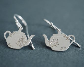 May include: A pair of silver dangle earrings featuring a teapot design with a floral pattern. The earrings are made of sterling silver and have a hook closure.