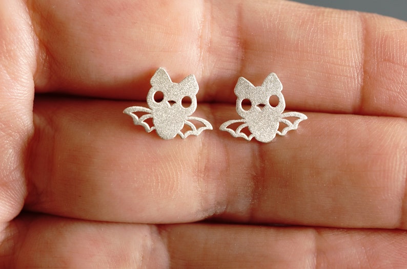 Peut inclure: Une paire de boucles d'oreilles en argent en forme de chauve-souris. Les chauves-souris ont un design mignon et cartoon avec de grands yeux.