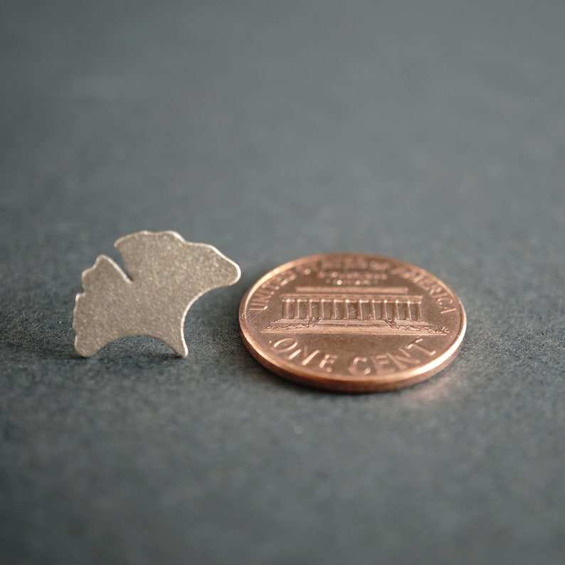 Op de afbeelding: Een zilverkleurige metalen uitsnijding van een ginkgo-blad, naast een Amerikaanse cent. De munt is koperkleurig en heeft het Lincoln Memorial en de woorden "United States of America" en "One Cent".