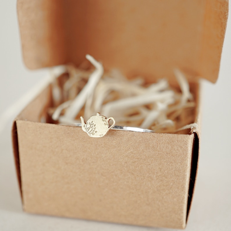 K&ouml;nnte beinhalten: Ein silbernes Armreif-Armband mit einem kleinen goldenen Teekannen-Anh&auml;nger. Der Anh&auml;nger hat ein detailliertes Design einer Teekanne mit einem floralen Muster.