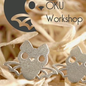 Peut inclure: Deux boucles d'oreilles en forme de chauve-souris argent&eacute;es avec des ailes et des yeux d&eacute;coup&eacute;s. Les boucles d'oreilles sont sur un lit de copeaux de bois. L'image comprend le texte "OKU Workshop" et "design & laser cut okuworkshop@hotmail.com".
