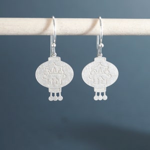 Può includere: Un paio di orecchini d'argento a forma di lanterne cinesi con un design delicato. Gli orecchini sono appesi a una superficie di legno bianco.