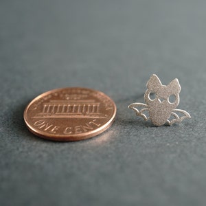 Peut inclure: Un charme en forme de chauve-souris de couleur argent&eacute;e, pr&eacute;sent&eacute; &agrave; c&ocirc;t&eacute; d'un centime am&eacute;ricain pour comparaison de taille. La chauve-souris a une silhouette d&eacute;taill&eacute;e avec les ailes d&eacute;ploy&eacute;es.