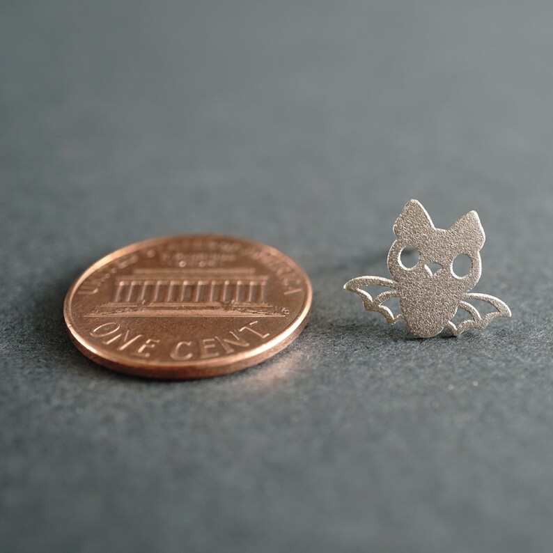 Peut inclure: Un charme en forme de chauve-souris de couleur argent&eacute;e, pr&eacute;sent&eacute; &agrave; c&ocirc;t&eacute; d'un centime am&eacute;ricain pour comparaison de taille. La chauve-souris a une silhouette d&eacute;taill&eacute;e avec les ailes d&eacute;ploy&eacute;es.