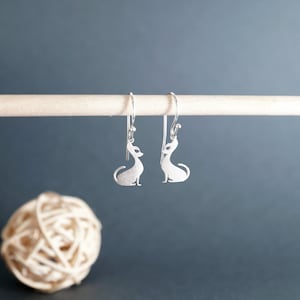 May include: A pair of silver dangle earrings featuring a silhouette of a cat. The earrings are hanging from a wooden dowel rod.
