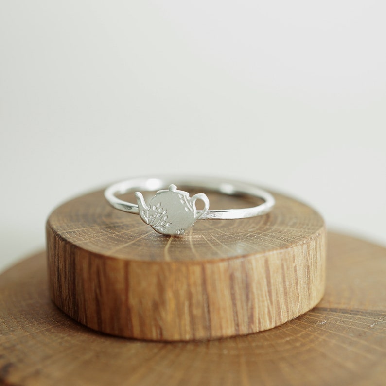 K&ouml;nnte beinhalten: Ein silberner Ring mit einem Teekannen-Design. Die Teekanne ist mit einem floralen Muster graviert. Der Ring liegt auf einer Holzoberfl&auml;che.