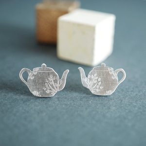 Puede incluir: Un par de pendientes de plata en forma de tetera con un diseño floral. Los pendientes son pequeños y delicados, y serían un gran regalo para un amante del té.