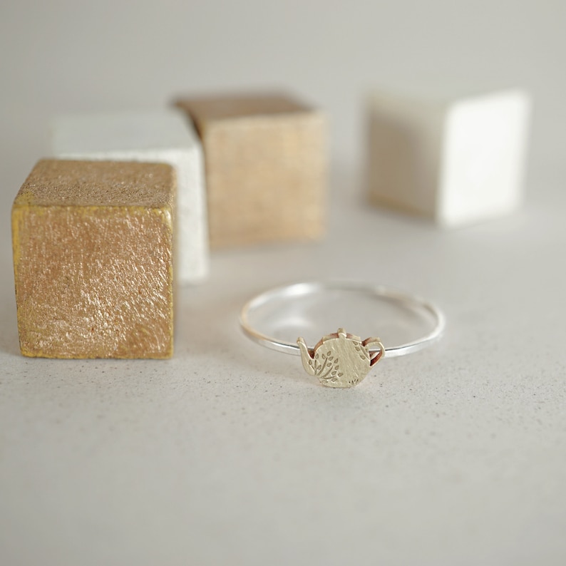 K&ouml;nnte beinhalten: Ein silberner Ring mit einem goldenen Teekannen-Anh&auml;nger. Die Teekanne hat ein florales Design.
