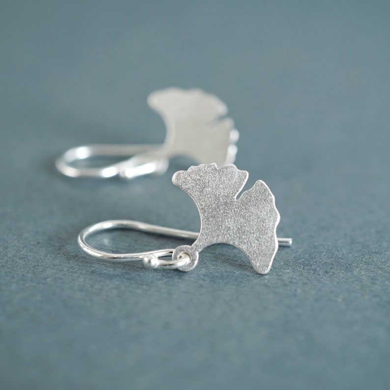 May include: A pair of silver earrings shaped like ginkgo leaves. The leaves have a textured surface and are attached to silver hooks.