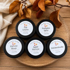 May include: Five round black metal candle tins with white labels. Each label reads The Nifty Candle, a scent name, fall sampler, and 1.5oz/42g. The scents are Fall Leaves, Pumpkin Spice, Harvest Moon, Apple Harvest, and Snickerdoodle.