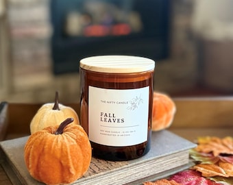 May include: A brown glass jar candle with a light wood lid and a white label reading THE NIFTY CANDLE FALL LEAVES SOY WAX CANDLE 10 OZ 283 G HANDCRAFTED IN ARIZONA, positioned next to two small orange fabric pumpkins on a book.