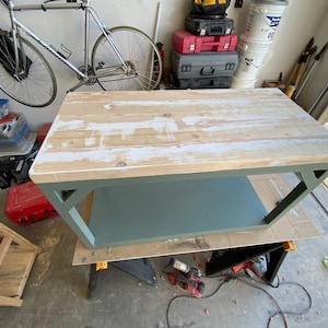 May include: A wooden table with a light wood top and a green painted base. The table has a shelf underneath the top. The table is in a garage setting.