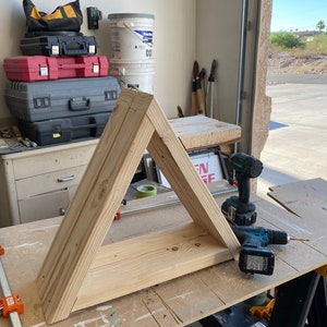 May include: A wooden triangle shelf is being assembled on a workbench. Two cordless drills are visible on the workbench. The shelf is made of unfinished wood.