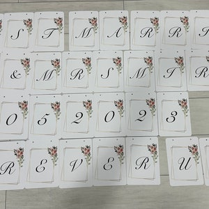 May include: A collection of white cards with black calligraphy letters and numbers. The cards spell out "Just Married" and "Forever Us" with the date "05 02 2023". Each card has a floral design and a gold border.