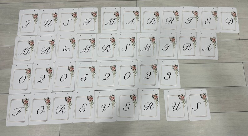 May include: A collection of white cards with black calligraphy letters and numbers. The cards spell out "Just Married" and "Forever Us" with the date "05 02 2023". Each card has a floral design and a gold border.