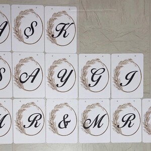 May include: White rectangular cards with black letters and a decorative floral wreath. The letters spell out names and the word "MRS & MRS". The cards are likely for a wedding or event.