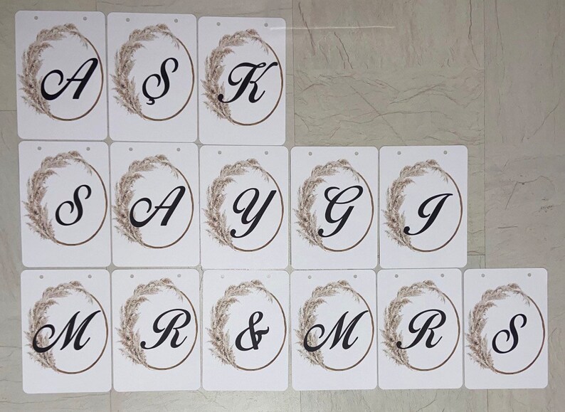 May include: White rectangular cards with black letters and a decorative floral wreath. The letters spell out names and the word "MRS & MRS". The cards are likely for a wedding or event.