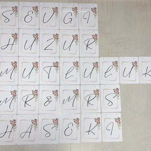May include: A collection of white cardstock banners with black cursive letters and floral accents. Each banner has a gold geometric border and a small hole at the top for hanging. The banners spell out phrases like "MR & MRS" and individual letters.