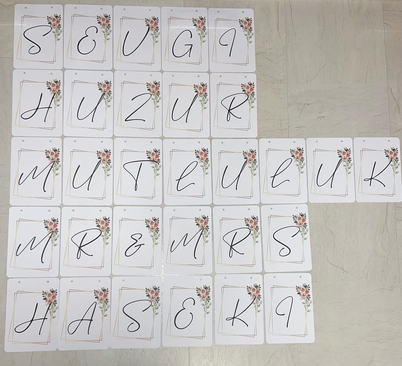 May include: A collection of white cardstock banners with black cursive letters and floral accents. Each banner has a gold geometric border and a small hole at the top for hanging. The banners spell out phrases like "MR & MRS" and individual letters.
