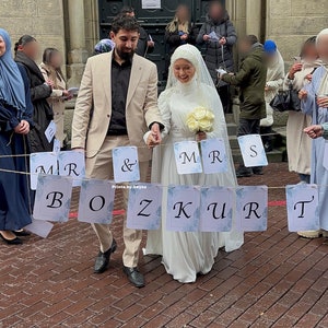 May include: A wedding scene featuring a couple walking together, holding hands. The bride wears a white dress and veil, carrying a bouquet. A banner with the words "Mr & Mrs BOZKURT" hangs in front of them. The groom wears a tan suit. The image includes the text "Prints.by.beyza".