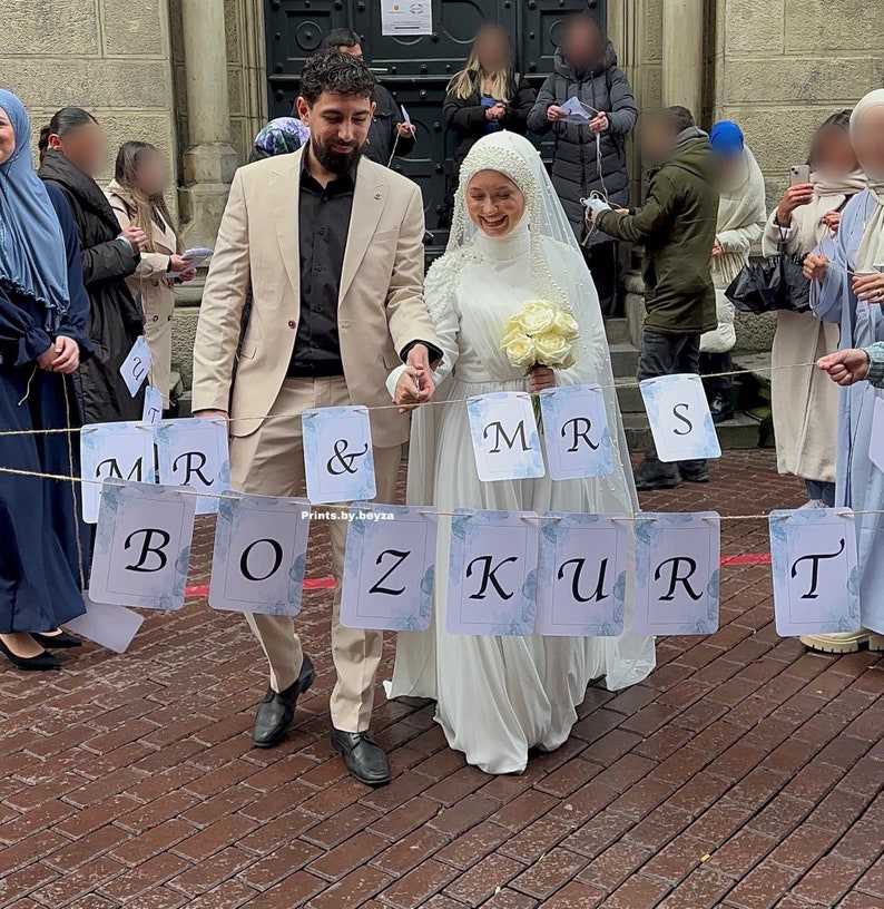 May include: A wedding scene featuring a couple walking together, holding hands. The bride wears a white dress and veil, carrying a bouquet. A banner with the words "Mr & Mrs BOZKURT" hangs in front of them. The groom wears a tan suit. The image includes the text "Prints.by.beyza".