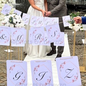 May include: Wedding banner with letters and floral accents. The banner features white rectangular cards with a gold geometric frame and pink floral details. The letters spell out a name and the word "LOVE". The banner is strung across a wedding ceremony.