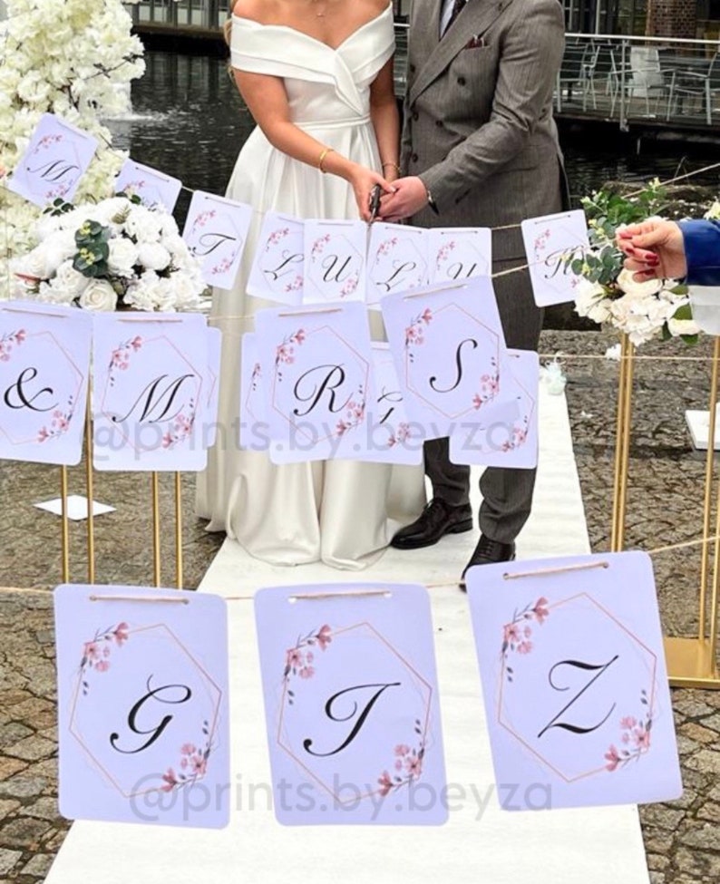 May include: Wedding banner with letters and floral accents. The banner features white rectangular cards with a gold geometric frame and pink floral details. The letters spell out a name and the word "LOVE". The banner is strung across a wedding ceremony.