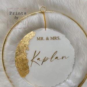 May include: A circular wedding sign with a gold-colored metal ring and chain. The sign is white with gold leaf accents and gold lettering that reads "MR. & MRS. Kaplan". The sign is hanging on a white, fluffy surface.