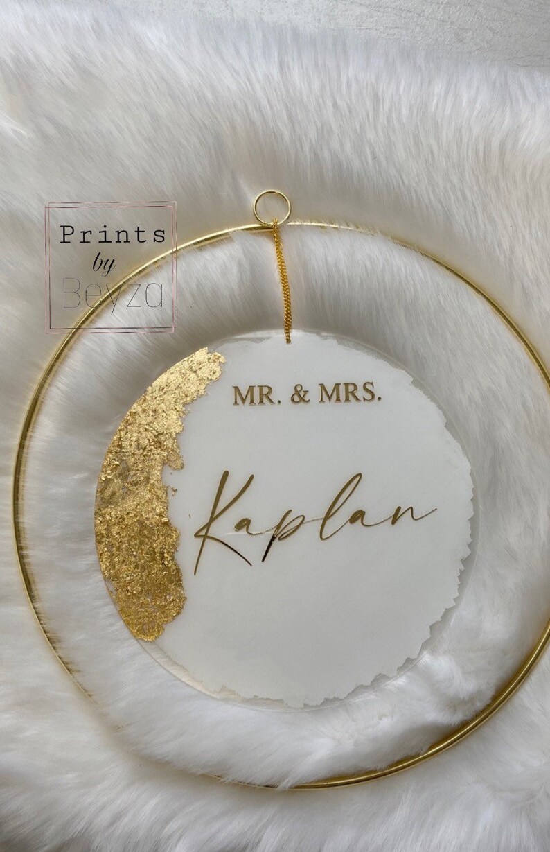 May include: A circular wedding sign with a gold-colored metal ring and chain. The sign is white with gold leaf accents and gold lettering that reads "MR. & MRS. Kaplan". The sign is hanging on a white, fluffy surface.