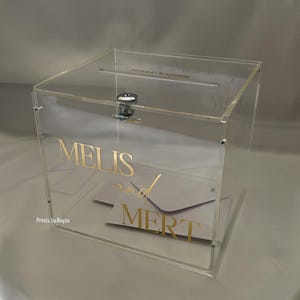 May include: Clear acrylic box with a slot on top, featuring the names "MELIS and MERT" in gold lettering. A silver latch secures the front. Inside, a white envelope is visible. The box is designed for events.