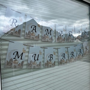 May include: A banner with the words "Ramadan Mubarak" printed on individual pennants. Each pennant features a mosque illustration with a crescent moon and lantern design. The letters are in a black, elegant font against a light blue background.