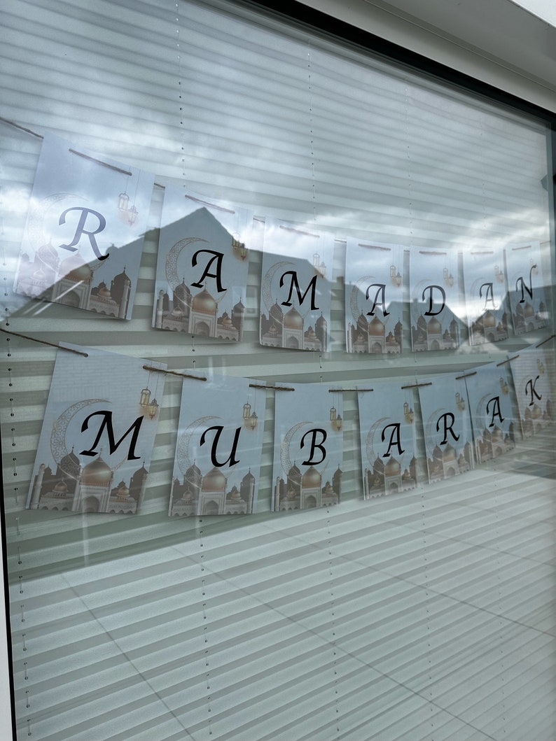 May include: A banner with the words "Ramadan Mubarak" printed on individual pennants. Each pennant features a mosque illustration with a crescent moon and lantern design. The letters are in a black, elegant font against a light blue background.