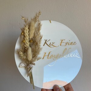 May include: A white, round decorative sign with gold script that reads "Kız Evine Hoşgeldiniz." The sign is adorned with a bundle of dried beige flowers and wheat tied with a ribbon. A small gold chain is attached for hanging.