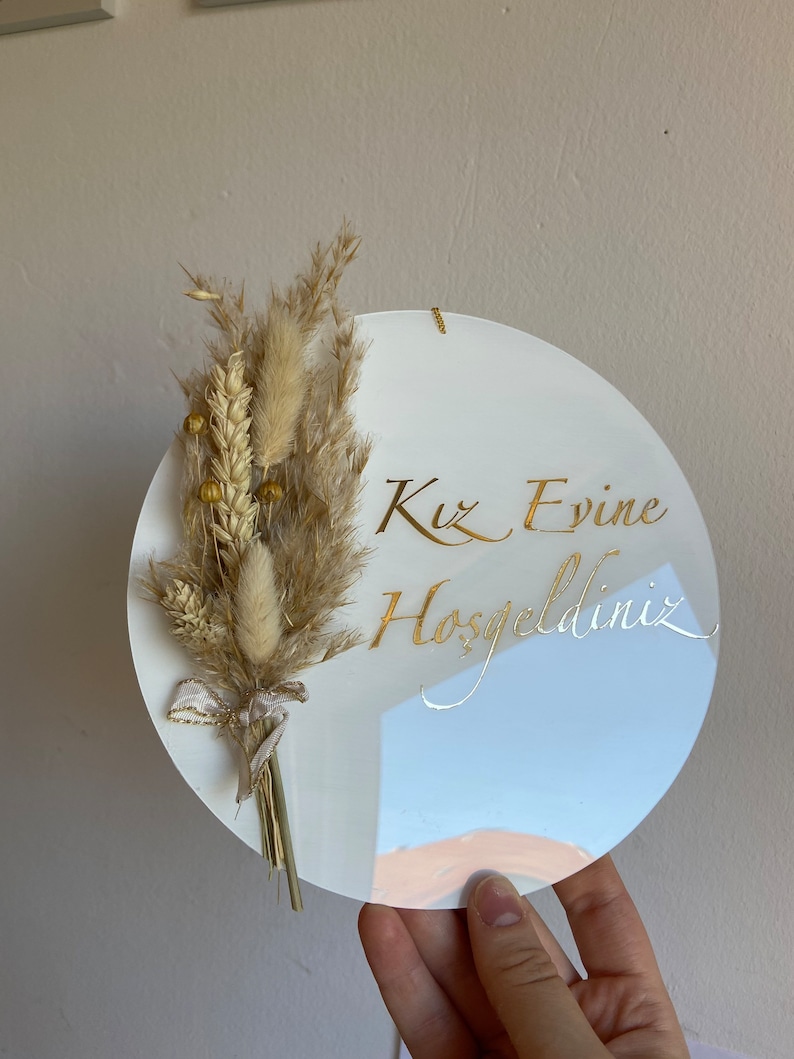 May include: A white, round decorative sign with gold script that reads "Kız Evine Hoşgeldiniz." The sign is adorned with a bundle of dried beige flowers and wheat tied with a ribbon. A small gold chain is attached for hanging.