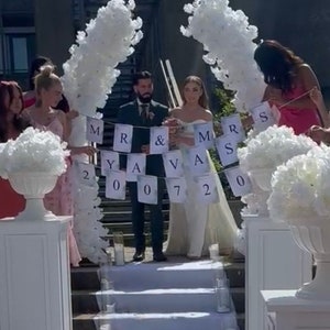 May include: A wedding ceremony scene with a couple standing under a floral arch. A banner reads "Mr & Mrs Yava 200720". White floral arrangements and a white carpet lead to the couple. People are standing on either side of the couple.