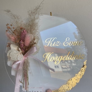 May include: A clear, round decorative sign with gold lettering that reads "Kız Evine Hoşgeldiniz". It features a dried flower arrangement with pink and beige tones, tied with a pink ribbon. Gold leaf accents the bottom edge.