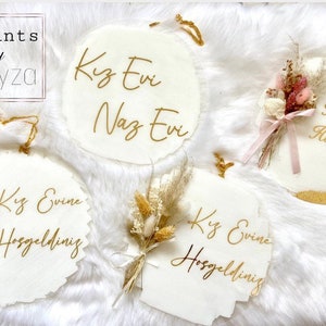May include: White, circular decorative signs with gold lettering and dried floral accents. The signs feature the text "Kiz Evine Hosgeldiniz" and "Kiz Evi Naz Evi". Some signs have a gold trim and are tied with gold string.