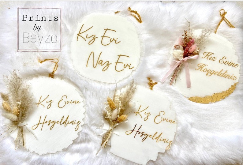 May include: White, circular decorative signs with gold lettering and dried floral accents. The signs feature the text "Kiz Evine Hosgeldiniz" and "Kiz Evi Naz Evi". Some signs have a gold trim and are tied with gold string.