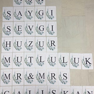 May include: A collection of white rectangular cards with black letters spelling out words. Each card features a gold border and a watercolor-style eucalyptus leaf design. The words include "ASK", "SAYGI", "SEVGI", "HUZUR", "MUTLULUK", "MR & MRS", and "&Ccedil;ALIŞKAN".