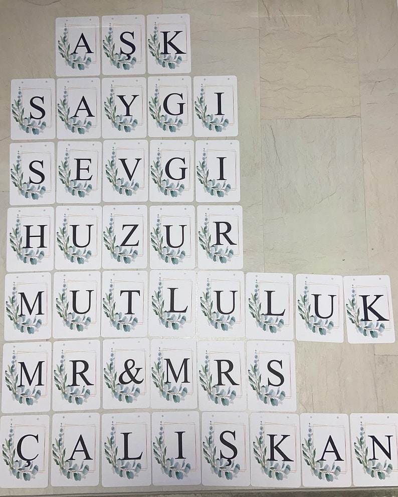 May include: A collection of white rectangular cards with black letters spelling out words. Each card features a gold border and a watercolor-style eucalyptus leaf design. The words include "ASK", "SAYGI", "SEVGI", "HUZUR", "MUTLULUK", "MR & MRS", and "&Ccedil;ALIŞKAN".