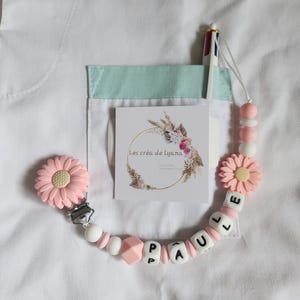 Peut inclure: Une attache-sucette personnalisée avec des perles en silicone en forme de marguerite rose pâle, des perles sphériques blanches et roses, et des cubes blancs formant le nom PAULLE. Elle inclut un clip métallique et une carte Les créa de Lyana.