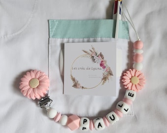 May include: A personalized pacifier clip featuring light pink silicone daisy beads, white and pink spherical beads, and white cube beads spelling the name PAULLE. The clip includes a metal fastener and a square card with the text Les créa de Lyana.