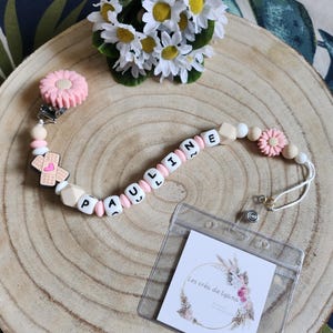Pode incluir: Um cordão de contas com silicone rosa e branco, um pendente em forma de penso e contas com letras brancas que formam o nome PAULINE. Inclui um clipe de flor rosa e um porta-cartão de plástico transparente com o texto Les créa de Lyana.